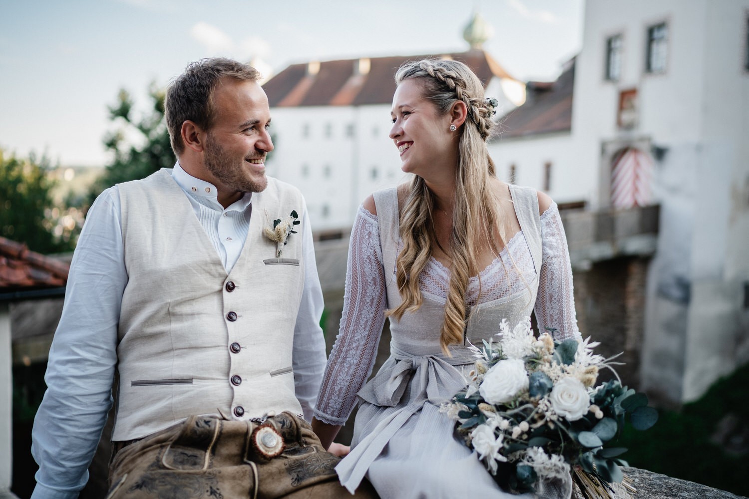 Hochzeit in der Veste Oberhaus Passau, Paarshooting, Brautpaar sitzt auf Mauer und lächelt sich an