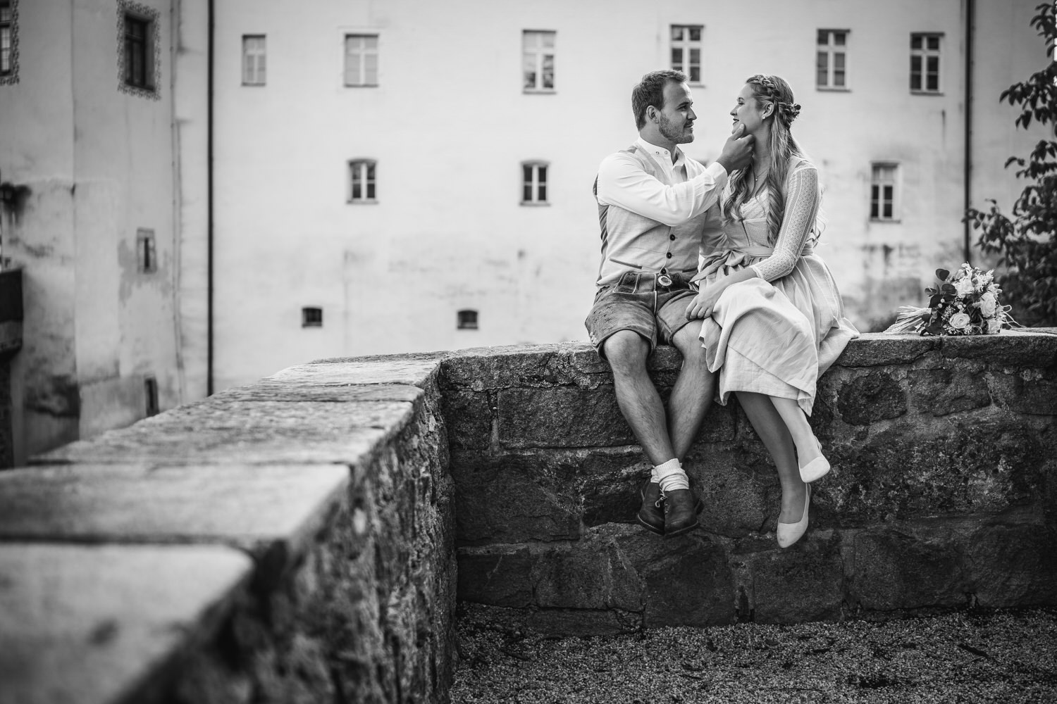 Hochzeit in der Veste Oberhaus Passau, Paarshooting, Brautpaar sitzt auf Mauer und streichelt sich über das Gesicht