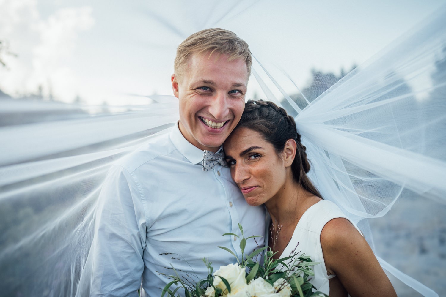 Hochzeitspaar lacht gemeinsam in die Kamera unter dem Schleier mit dem Brautstrauss in der Hand