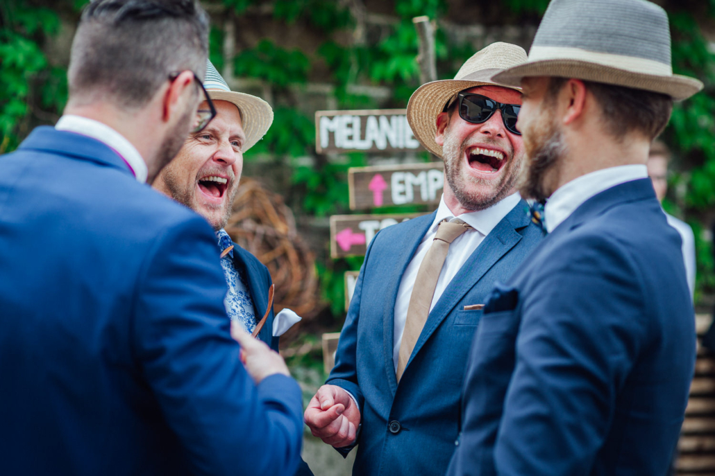Hochzeitsgäste in Anzügen und Strohhüten lachen gemeinsam bei der freien Trauung auf Gut Aichet