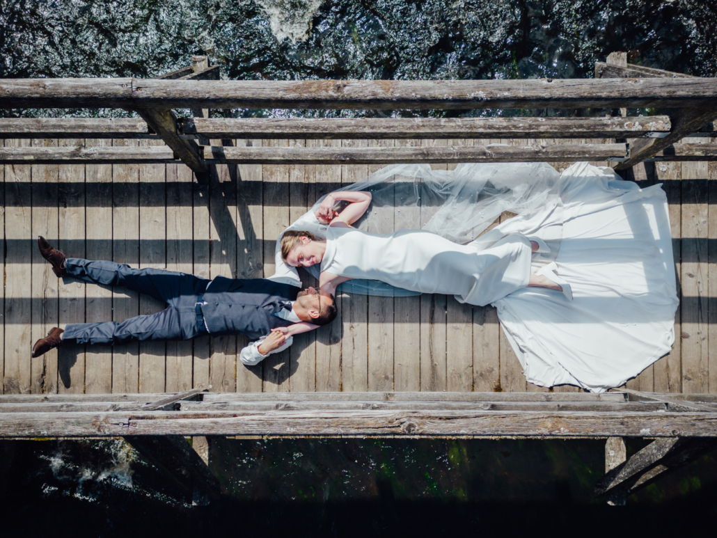 Hochzeitspaar liegt entspannt auf einer Holzbrücke, aufgenommen aus der Vogelperspektive.